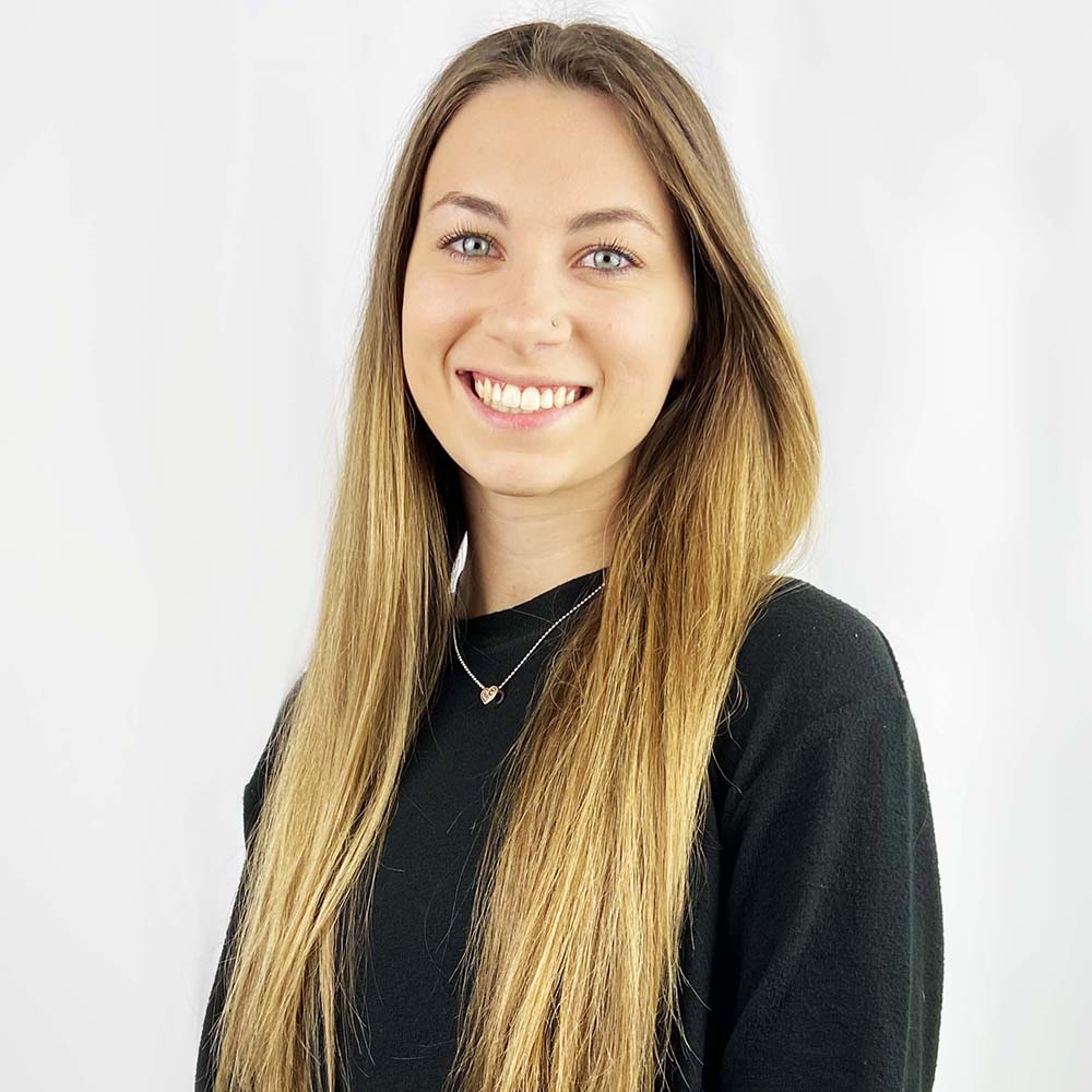 Smiling woman with long blonde hair wearing a black top and a heart-shaped pendant necklace, against a white background.