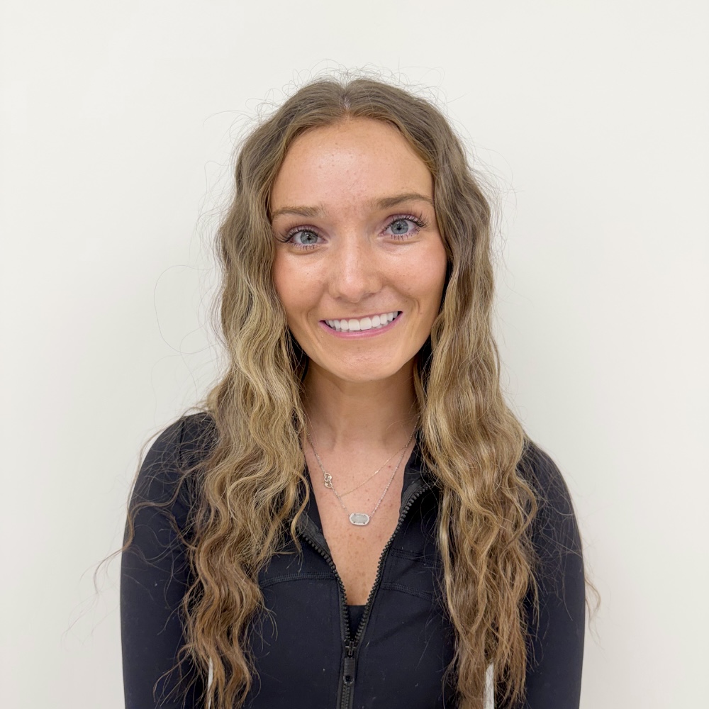 Portrait of a smiling woman with long wavy brown hair wearing a black zip-up jacket against a pale background.