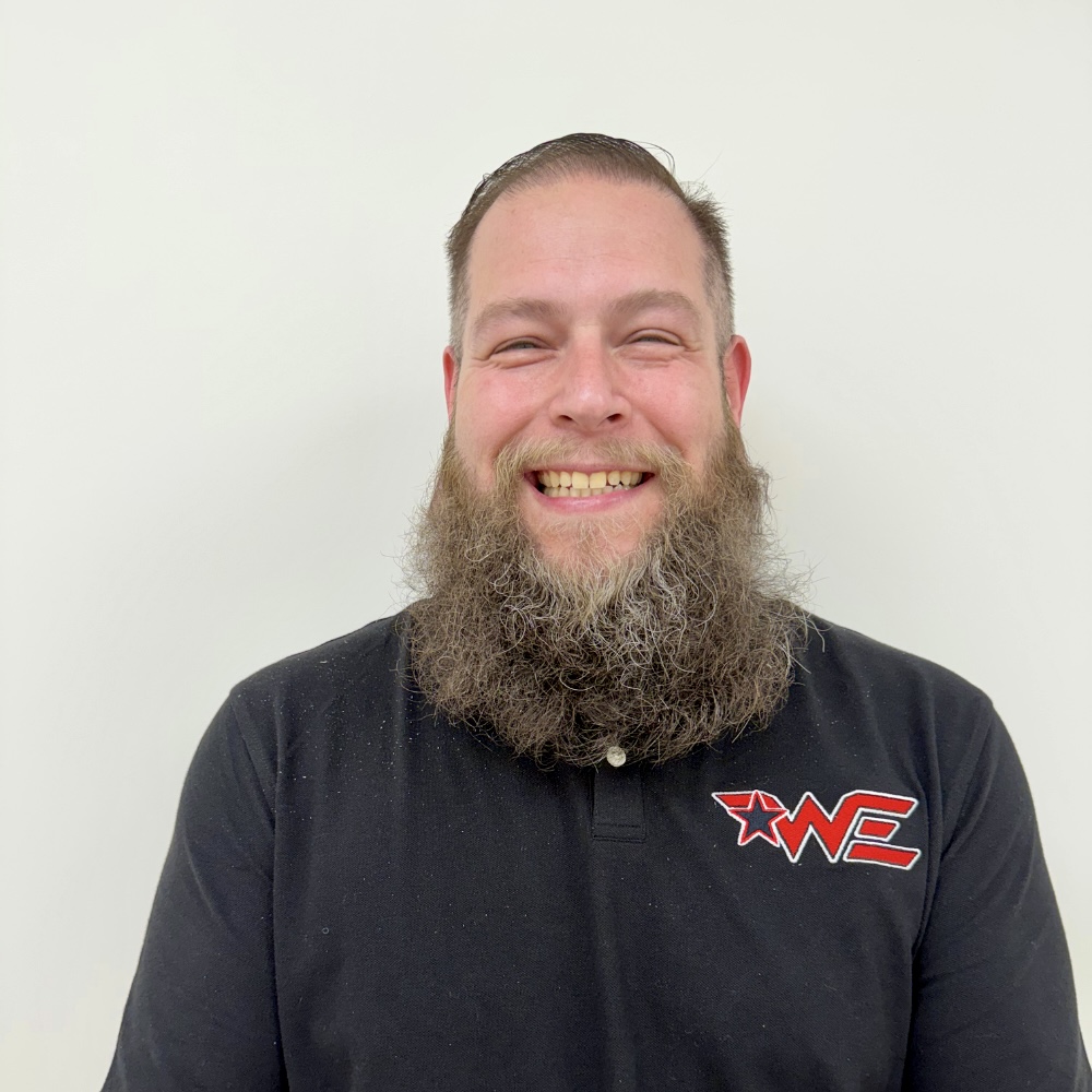 Smiling man with a thick beard and short hair wearing a black polo shirt with a red WE logo against a plain light background.