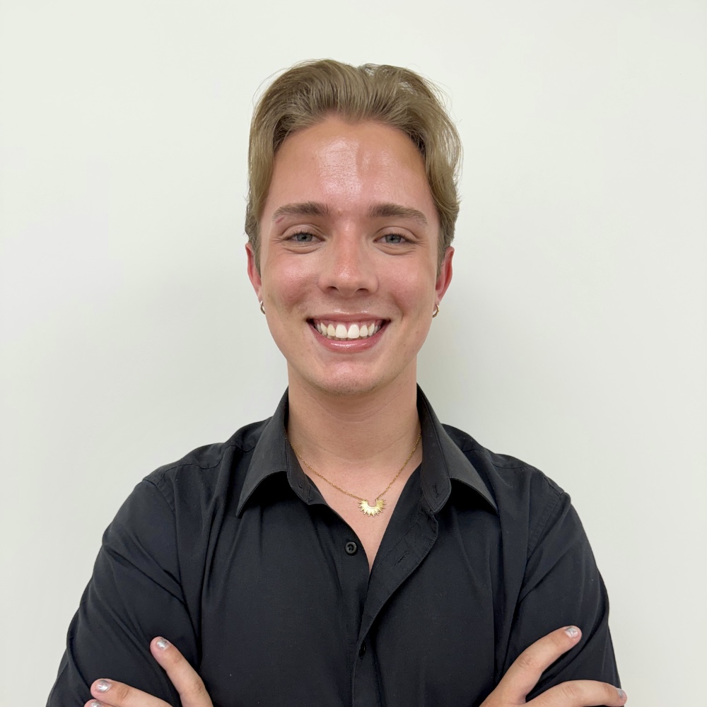 Smiling person with short blond hair wearing a black button-down shirt, arms crossed, against a light wall.