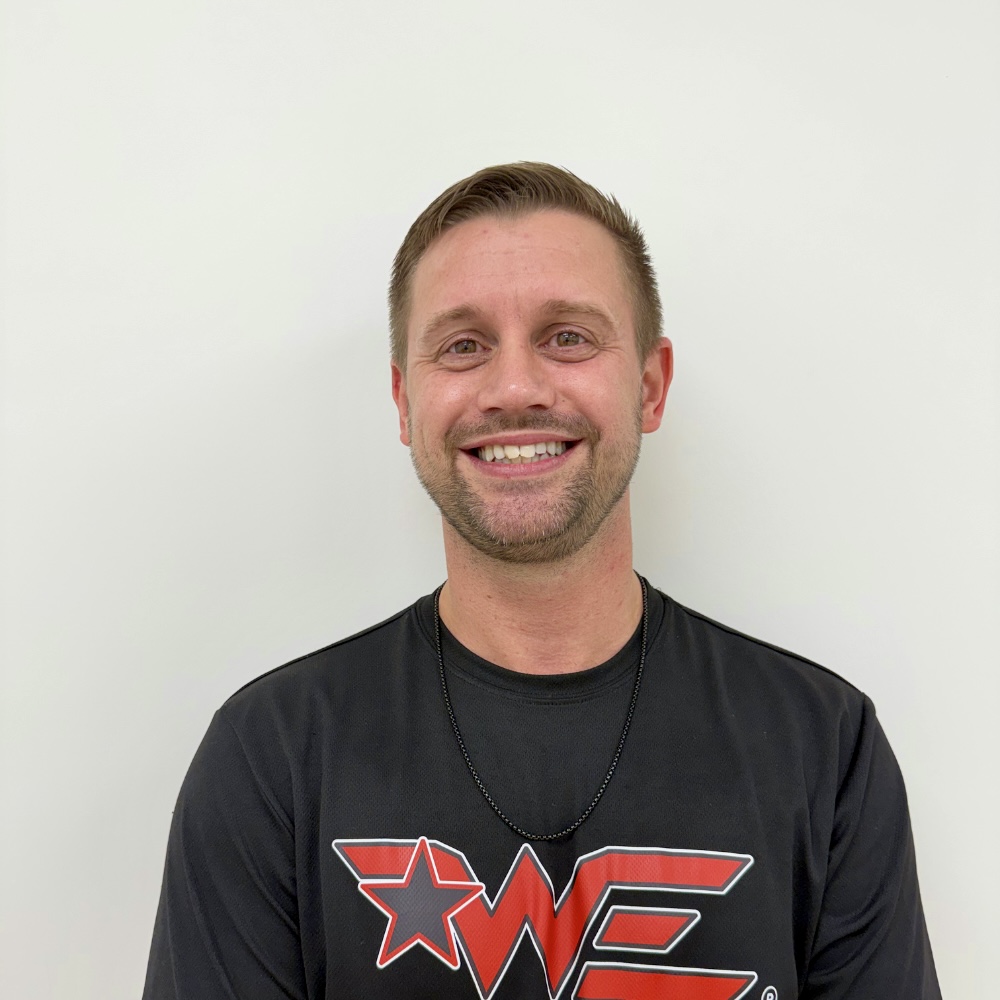 Smiling man with short light brown hair in a black t-shirt with a red star and 'WE' logo, against a light background.